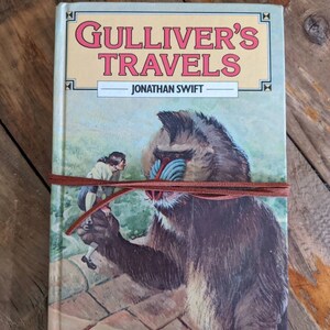May include: A vintage hardcover book titled "Gulliver's Travels" by Jonathan Swift. The cover features an illustration of a large, brown, furry creature with a blue face and a small human figure standing on a stone surface.