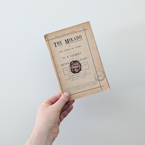 May include: A vintage programme for a performance of "The Mikado" by W.S. Gilbert and Arthur Sullivan. The programme is printed on aged paper and features a red seal with the words "Chappell & Co. Ltd."