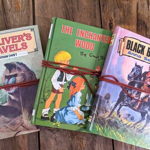 May include: Three vintage hardcover books with brown leather straps wrapped around them. The books are titled "Gulliver's Travels", "The Enchanted Wood", and "Black Beauty".
