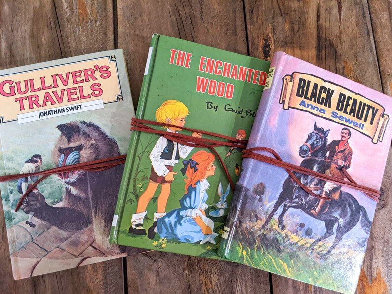 May include: Three vintage hardcover books with brown leather straps wrapped around them. The books are titled "Gulliver's Travels", "The Enchanted Wood", and "Black Beauty".