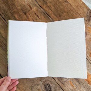 May include: A blank, white, paper notebook with a textured cover. The notebook is open to a blank page and is lying on a wooden surface.