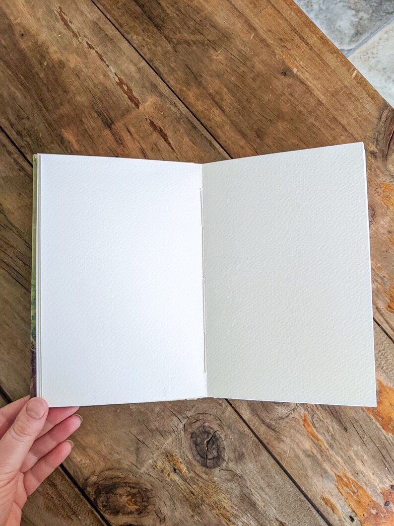 May include: A blank, white, paper notebook with a textured cover. The notebook is open to a blank page and is lying on a wooden surface.