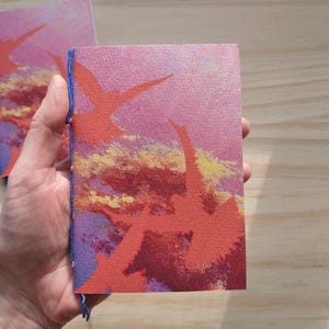 May include: A small rectangular notebook with a textured cover featuring a printed design of orange birds flying against a pink, purple, and yellow sunset sky. The notebook is bound along the left edge with visible blue thread stitching.