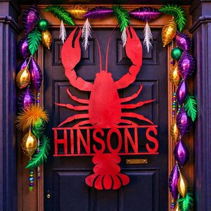 May include: A red crawfish door decoration with the name "HINSON'S" cut out below. The door is decorated with purple, gold, and green Mardi Gras beads and accents. Feathers and decorative leaves are also included.
