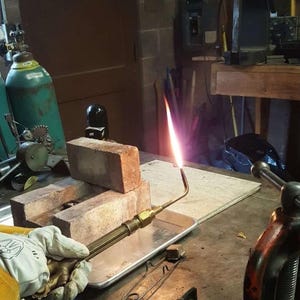 May include: A welding torch with a bright flame is held over a metal tray. The torch is aimed at a stack of red bricks. A gloved hand holds the torch. A green gas cylinder is in the background.