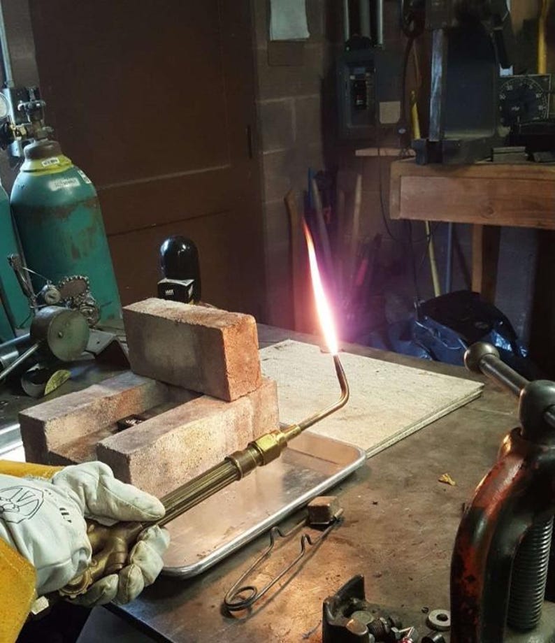 May include: A welding torch with a bright flame is held over a metal tray. The torch is aimed at a stack of red bricks. A gloved hand holds the torch. A green gas cylinder is in the background.