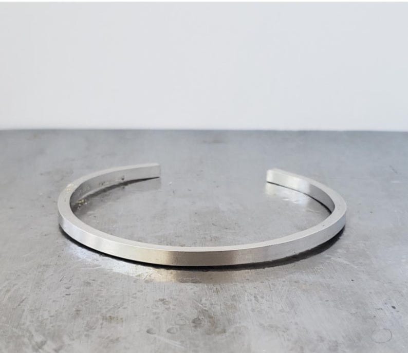 May include: A silver cuff bracelet with a flat, rectangular profile. The open-ended bracelet is made of a shiny metal and rests on a metallic surface. The minimalist design is suitable for everyday wear.