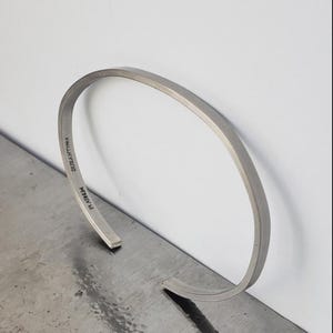 May include: A silver-coloured, open cuff bracelet. The bracelet has a flat, rectangular profile with rounded edges. The text "DUZANTINO" and "RAINM" are engraved on the inside. The bracelet is displayed on a metallic surface.