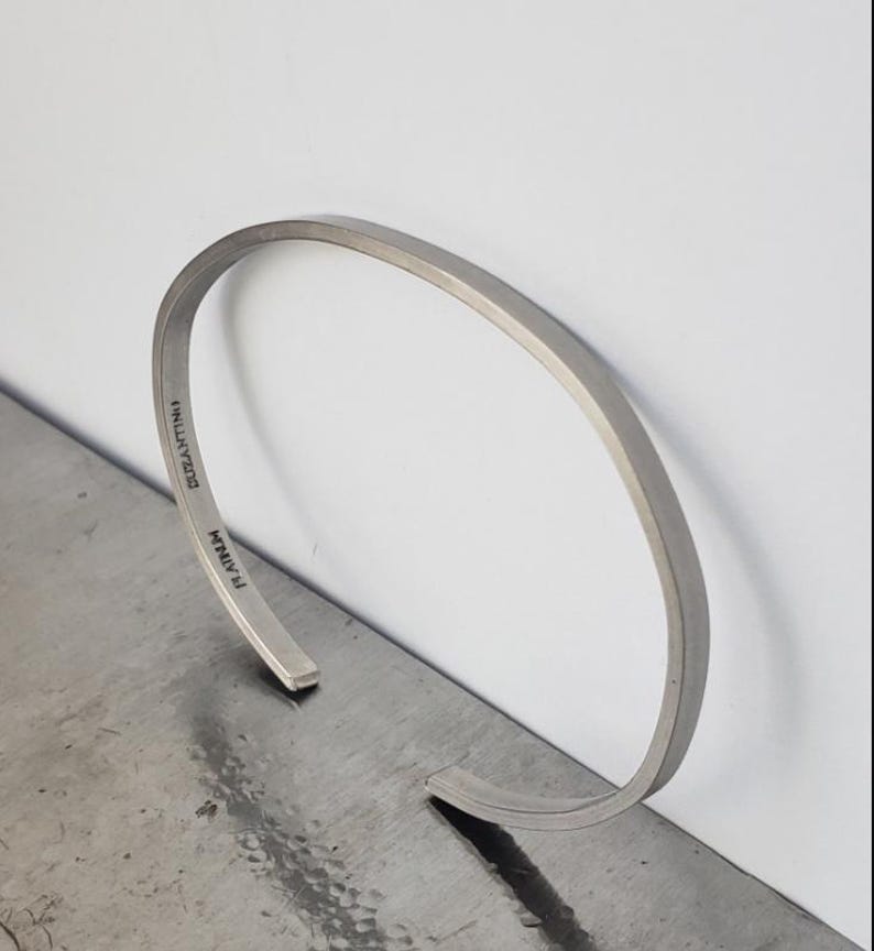 May include: A silver-coloured, open cuff bracelet. The bracelet has a flat, rectangular profile with rounded edges. The text "DUZANTINO" and "RAINM" are engraved on the inside. The bracelet is displayed on a metallic surface.