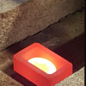 May include: A rectangular, red-orange container with a glowing, yellow-white interior. The container is set within a rough, textured, tan-coloured surface, creating a contrast of colour and texture.