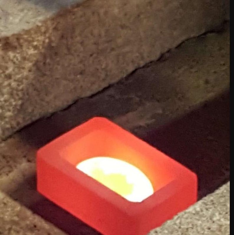 May include: A rectangular, red-orange container with a glowing, yellow-white interior. The container is set within a rough, textured, tan-coloured surface, creating a contrast of colour and texture.