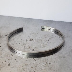 May include: A silver cuff bracelet with a flat, rectangular profile. The open-ended design features a subtle curve, and the metal has a brushed finish. The word "RAIN" is engraved on the inside of the bracelet.