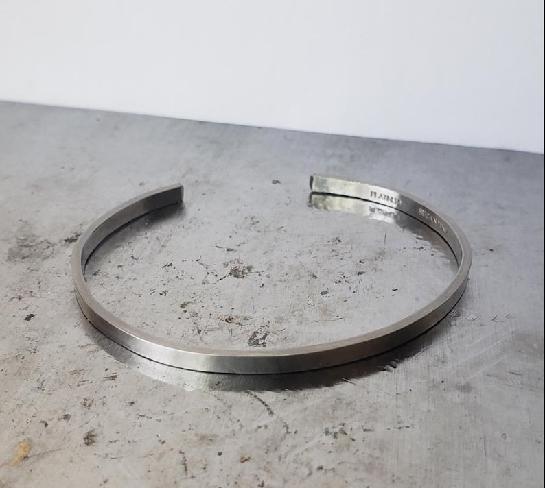 May include: A silver cuff bracelet with a flat, rectangular profile. The open-ended design features a subtle curve, and the metal has a brushed finish. The word "RAIN" is engraved on the inside of the bracelet.