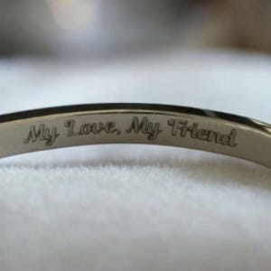 May include: A silver bangle bracelet with the inscription "My Love, My Friend". The bracelet has a polished finish and a simple design. The text is engraved in a delicate, cursive font. A piece of jewellery.