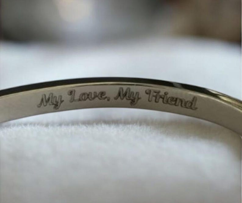 May include: A silver bangle bracelet with the inscription "My Love, My Friend". The bracelet has a polished finish and a simple design. The text is engraved in a delicate, cursive font. A piece of jewellery.