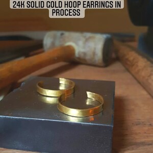 May include: Two gold hoop earrings on a black surface. The text "24K SOLID GOLD HOOP EARRINGS IN PROCESS" is above the earrings.