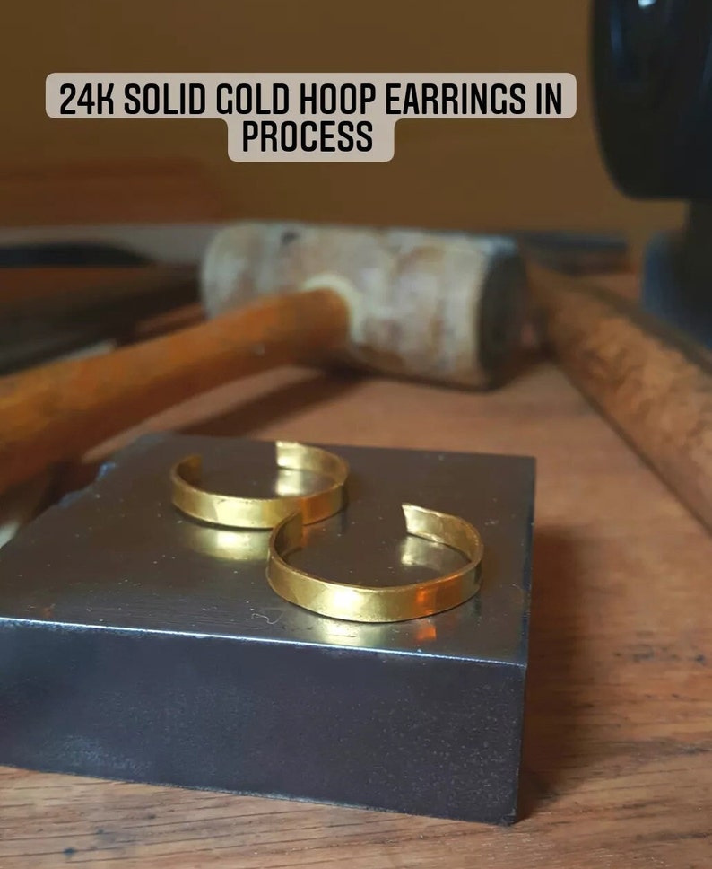May include: Two gold hoop earrings on a black surface. The text "24K SOLID GOLD HOOP EARRINGS IN PROCESS" is above the earrings.