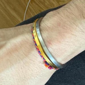 May include: A wrist displaying a silver bangle, a gold bangle, and a vibrant woven bracelet. The bracelets are stacked, creating a layered effect. The woven bracelet incorporates pink, orange, and blue threads.