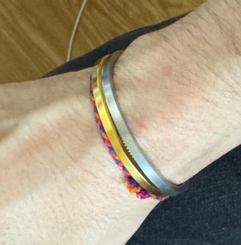 May include: A wrist displaying a silver bangle, a gold bangle, and a vibrant woven bracelet. The bracelets are stacked, creating a layered effect. The woven bracelet incorporates pink, orange, and blue threads.