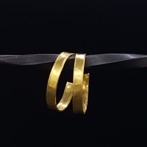 May include: Two gold-coloured hoop earrings, each with a hammered texture, are stacked on top of each other. The earrings are displayed against a black background.