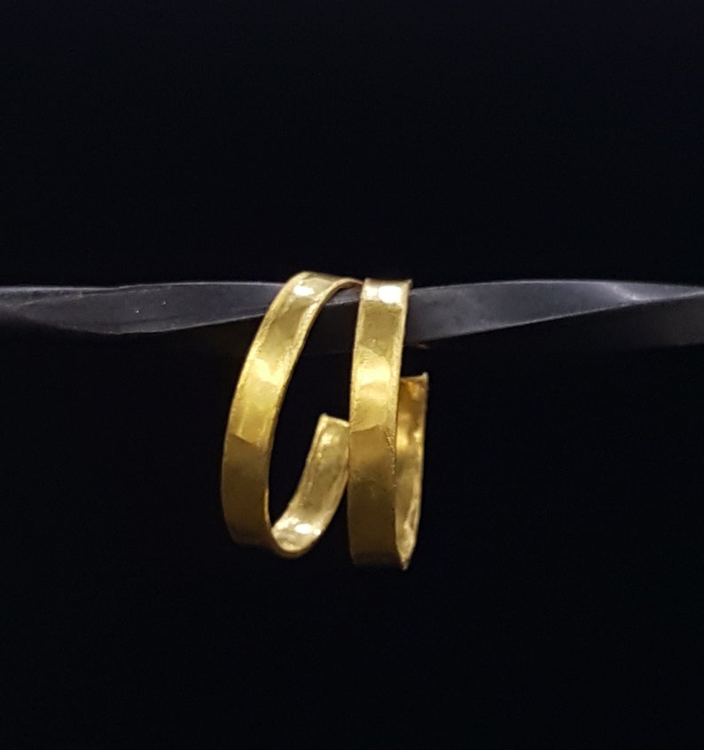 May include: Two gold-coloured hoop earrings, each with a hammered texture, are stacked on top of each other. The earrings are displayed against a black background.