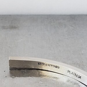 May include: A curved, silver-coloured metal object with the word "SUZANTINO" engraved on the top surface. The word "PLATINUM" is also engraved on the object. The object is set against a grey background.