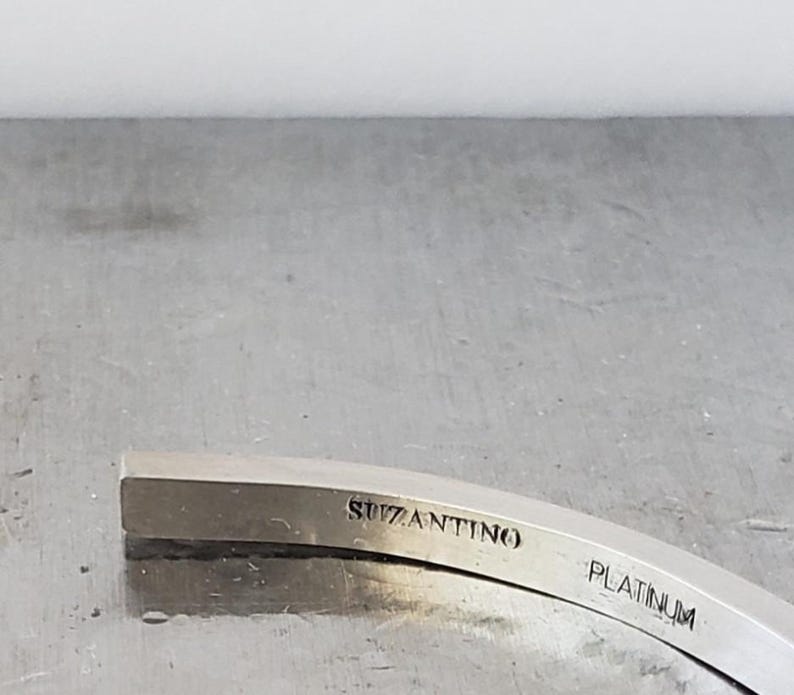 May include: A curved, silver-coloured metal object with the word "SUZANTINO" engraved on the top surface. The word "PLATINUM" is also engraved on the object. The object is set against a grey background.