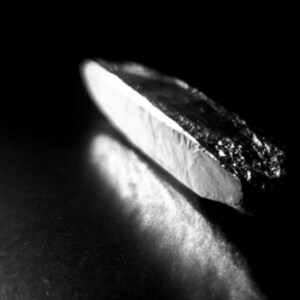 May include: A monochrome image showing a small, angular object with a bright, white edge and a dark, textured surface. The object is set against a dark, blurred background, creating a high-contrast effect.