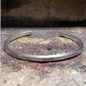 May include: A silver cuff bracelet with a flat, rectangular design and a brushed finish. The bracelet is slightly curved and open at the ends. The text "STANTIVM. R" is engraved on the inside. The bracelet is displayed on a textured surface.