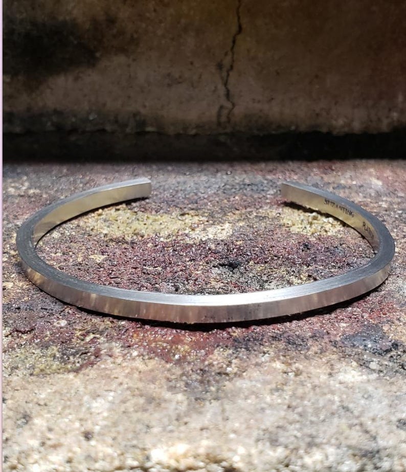 May include: A silver cuff bracelet with a flat, rectangular design and a brushed finish. The bracelet is slightly curved and open at the ends. The text "STANTIVM. R" is engraved on the inside. The bracelet is displayed on a textured surface.