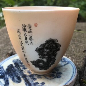 May include: A white ceramic teacup with black ink calligraphy and a small red seal. The calligraphy reads "愛一茶  葉  石  觀  山, 自  楽", which translates to "Love one tea, leaves, stone, watch the mountain, self-joy".