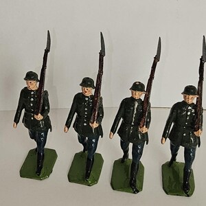 May include: Four vintage toy soldiers in a marching pose. Each soldier is dressed in a dark green uniform with a black helmet and holds a brown rifle with a silver bayonet. They stand on green rectangular bases.