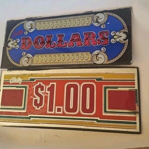 May include: Two vintage Bally slot machine signs. The top sign reads "DOLLARS" in red letters on a blue background, framed with gold accents. The bottom sign displays "$1.00" in red on a red and yellow background.
