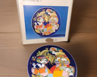 May include: A Rosenthal "Orientalische Nachtmusik" Wandteller Nr. 1 plate with a box. The plate is dark blue with a colourful illustration of figures in a patchwork design. The box has text in German.