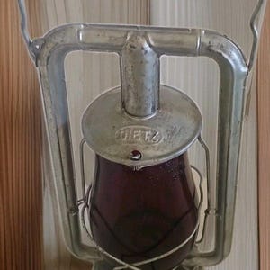 May include: Vintage Dietz lantern with a burgundy glass globe and a silver metal frame. The lantern has a handle for carrying and is designed to be hung. The word "DIETZ" is visible on the top. The lantern is likely used for camping or emergency lighting.
