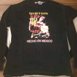 以下が含まれることがあります： ギターを弾いている人のグラフィックがプリントされた黒い長袖Tシャツ。シャツのテキストは「Para que se escuche la neta vota asi」と「HECHO EN MEXICO」です。