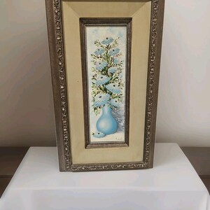 May include: A framed painting featuring a vase of blue flowers. The vase is a light blue color and the flowers are a lighter shade of blue with white accents. The frame is a dark, ornate design with a cream-colored matting.