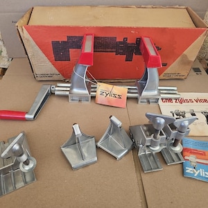 May include: A Zyliss vice set featuring a silver metal clamp with a red handle, several additional metal mounting brackets, and various components. The set includes a cardboard box with red and black graphics, a manual, and a product tag.