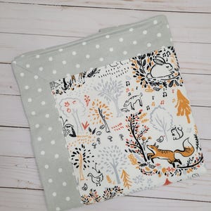 May include: A baby blanket with a woodland animal print. The blanket features a white background with illustrations of foxes, rabbits, and trees in orange, black, and gray. The border is a light gray with white polka dots.
