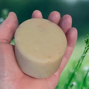 May include: A round, off-white bar of soap in a hand. The soap is smooth and has a slightly textured surface. The background is blurred and shows green grass and white flowers.