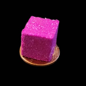 May include: A small, bright pink cube with a textured surface. The cube is sitting on a gold coin.