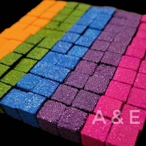 May include: A close-up of a collection of colorful, square-shaped objects arranged in diagonal rows. The colors include orange, green, blue, purple, and pink. The objects have a textured, glittery appearance. The letters "A & E" are visible in the bottom right corner.