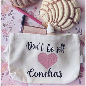 May include: Two concha pastries, one light brown and one beige, are on a floral background. A white canvas makeup bag with the text "Don't be self Conchas" and a heart-shaped concha graphic is in the foreground.