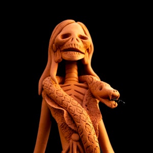 Op de afbeelding: Een terracotta beeld van een skeletvrouw die een jurk draagt met een slang eromheen gewikkeld. Het beeld is gedetailleerd met bloemen en ingewikkelde ontwerpen.