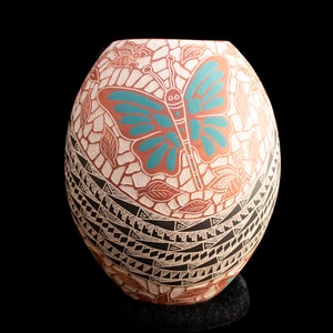 Puede incluir: Un jarrón de cerámica pintado a mano con un diseño de mariposa. El jarrón presenta una mariposa turquesa sobre un fondo terracota, rodeada de patrones de hojas y flores. La base tiene un diseño geométrico en blanco y negro.