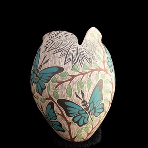 Puede incluir: Un jarrón de cerámica blanca con forma de corazón y un diseño geométrico. El jarrón está decorado con mariposas azules y verdes y hojas.