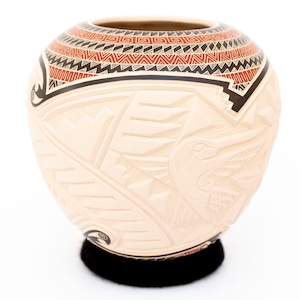Puede incluir: Una olla de cerámica blanca con un diseño tallado de un pájaro y patrones geométricos. La olla tiene un borde negro y está decorada con patrones geométricos rojos y negros.