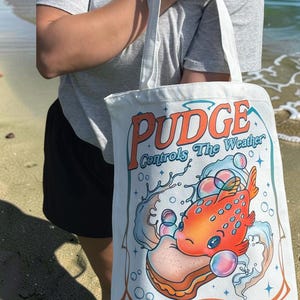May include: A white canvas tote bag featuring a colorful graphic of an orange fish holding a sandwich, surrounded by bubbles and water splashes. The text "PUDGE Controls The Weather" is printed in large orange and blue letters above the illustration.