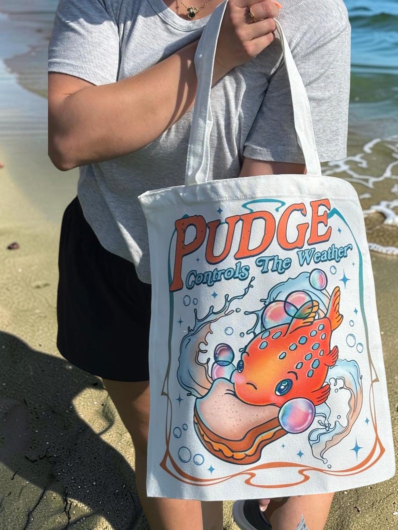 May include: A white canvas tote bag featuring a colorful graphic of an orange fish holding a sandwich, surrounded by bubbles and water splashes. The text "PUDGE Controls The Weather" is printed in large orange and blue letters above the illustration.