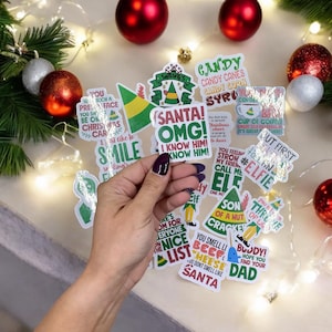 May include: A collection of colorful Christmas-themed stickers with quotes from the movie Elf. The stickers feature phrases like "Santa! OMG! I know him!" and "You smell like beef and cheese." Red and silver ornaments and Christmas lights are in the background.